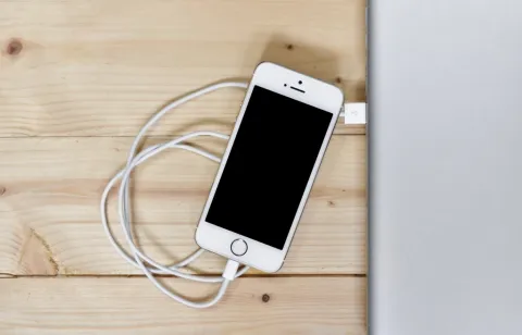 An iPhone resting on a wooden desk while connected to a charging cable.