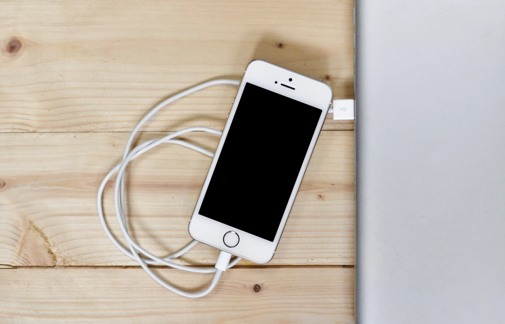 An iPhone resting on a wooden desk while connected to a charging cable.